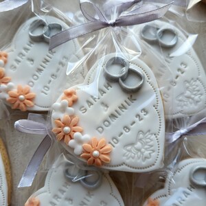 May include: White heart-shaped cookies with peach-colored flowers and silver rings. The cookies are decorated with the text "SARAH & DOMINIK 10-8-23".
