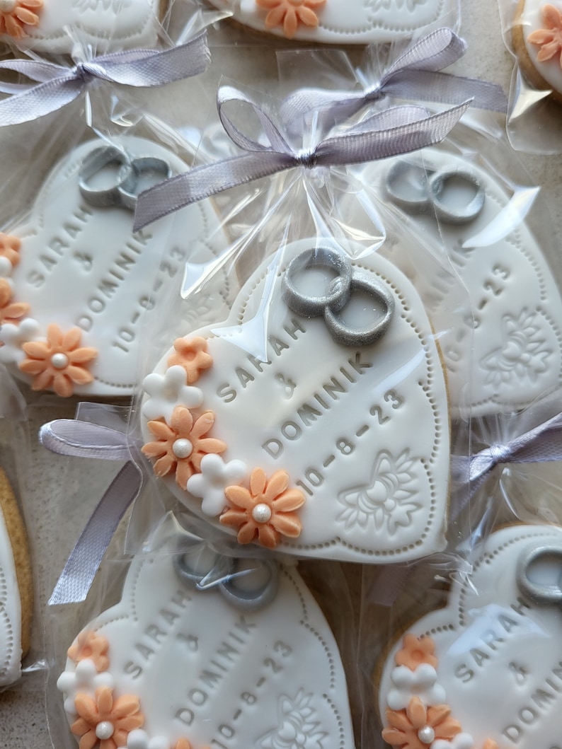 May include: White heart-shaped cookies with peach-colored flowers and silver rings. The cookies are decorated with the text "SARAH & DOMINIK 10-8-23".