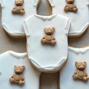 May include: White onesie-shaped cookies with a light brown teddy bear decoration. The cookies have a white icing base and a light brown border. The teddy bears are in a sitting position. These cookies are perfect for a baby shower.