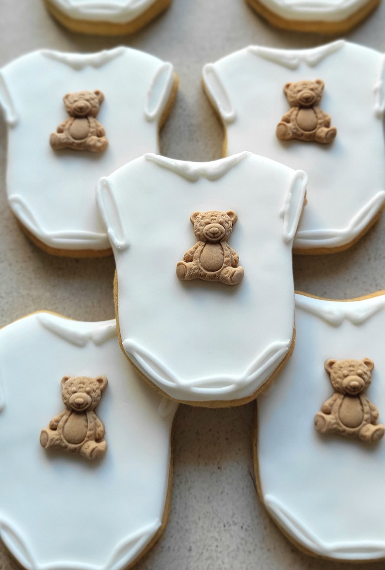May include: White onesie-shaped cookies with a light brown teddy bear decoration. The cookies have a white icing base and a light brown border. The teddy bears are in a sitting position. These cookies are perfect for a baby shower.