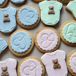 May include: Assortment of baby-themed cookies. Some are shaped like onesies with teddy bear decorations, others are round with heart designs. Colors include blue, pink, and green, with a light brown cookie base.