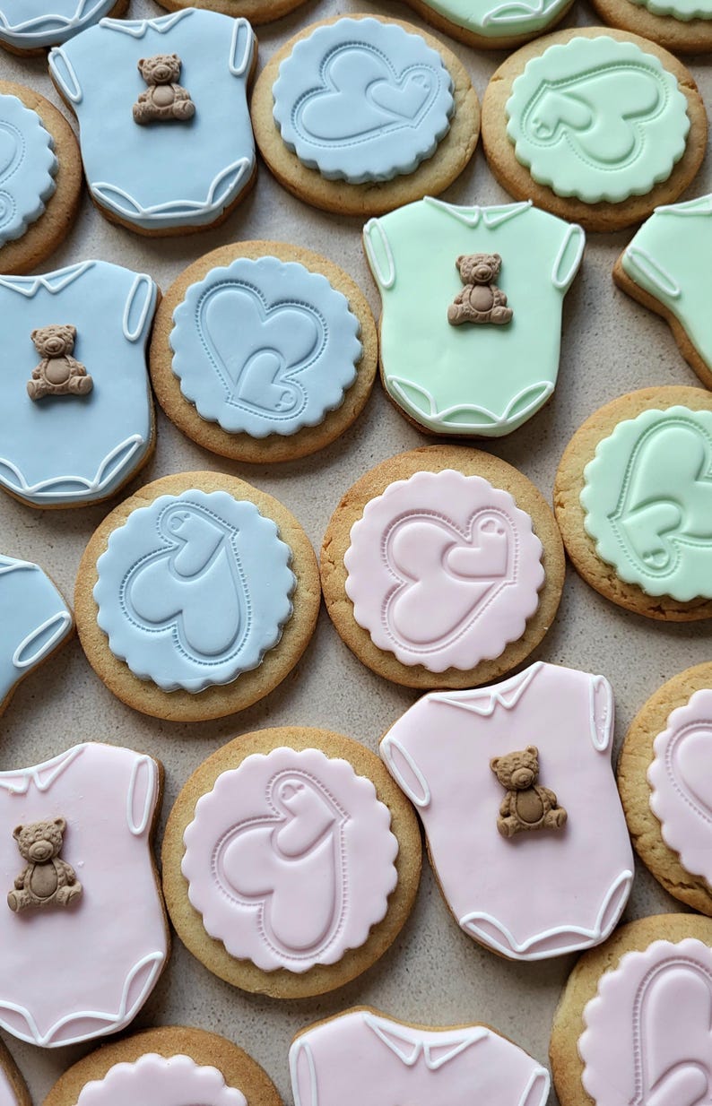 May include: Assortment of baby-themed cookies. Some are shaped like onesies with teddy bear decorations, others are round with heart designs. Colors include blue, pink, and green, with a light brown cookie base.