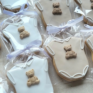 May include: Cookies shaped like baby onesies, decorated with a teddy bear and white icing. Each cookie is individually wrapped in clear cellophane and tied with a light purple ribbon. A sweet treat for a baby shower or newborn celebration.