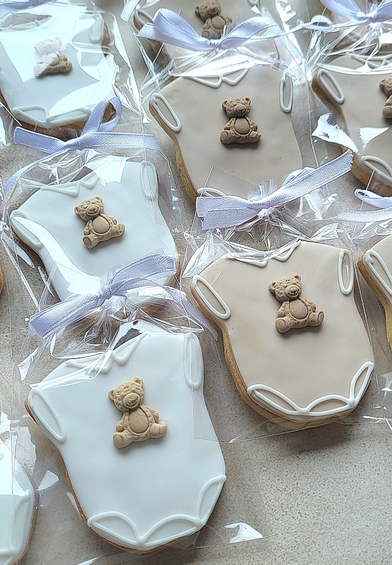 May include: Cookies shaped like baby onesies, decorated with a teddy bear and white icing. Each cookie is individually wrapped in clear cellophane and tied with a light purple ribbon. A sweet treat for a baby shower or newborn celebration.