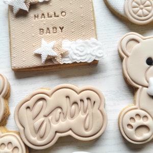 May include: A set of six decorated sugar cookies with a baby theme. The cookies are decorated with brown and white icing and feature designs such as a teddy bear, a baby carriage, and the word "Baby". One cookie has the text "Hallo Baby" in brown icing.