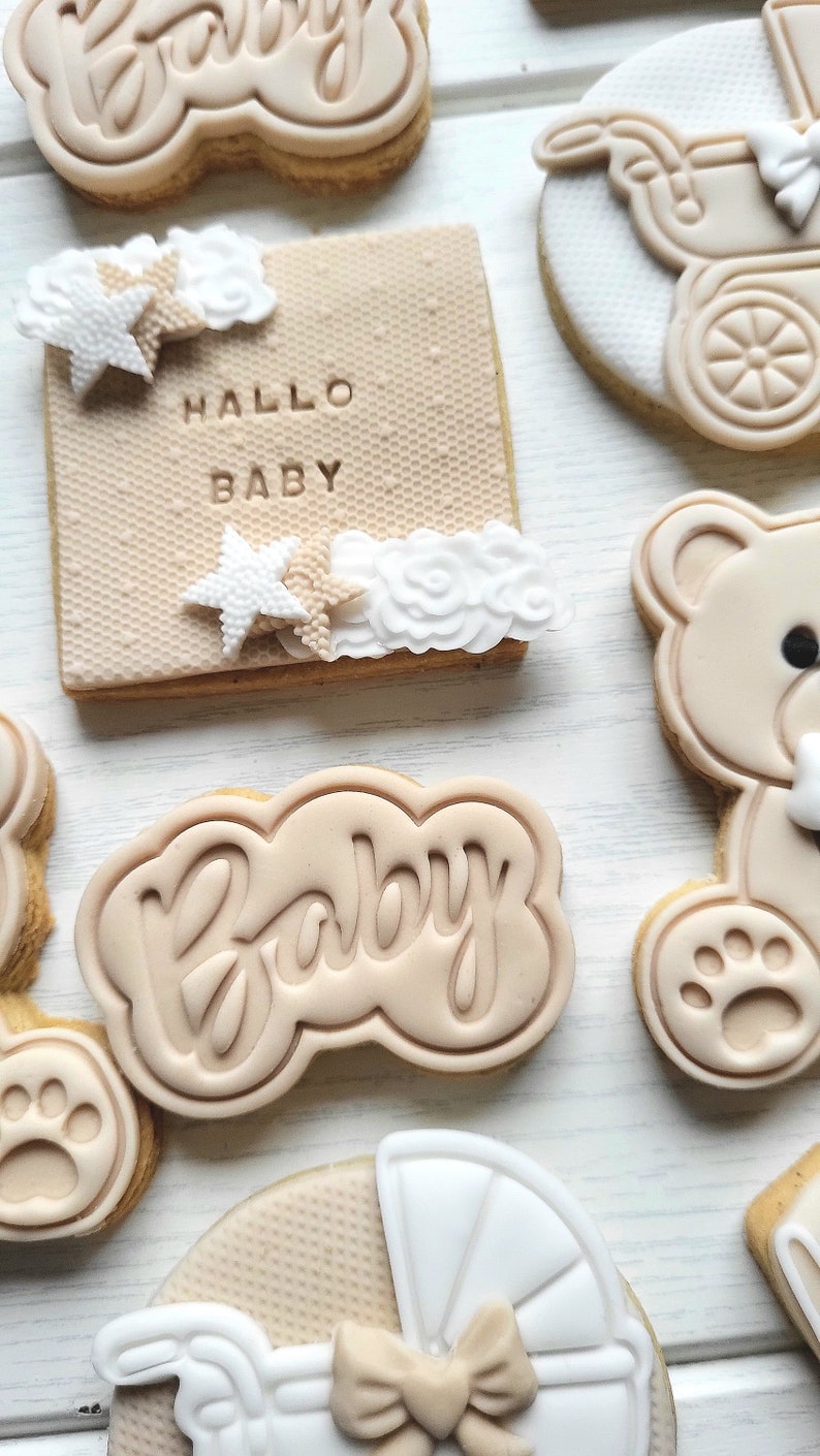 May include: A set of six decorated sugar cookies with a baby theme. The cookies are decorated with brown and white icing and feature designs such as a teddy bear, a baby carriage, and the word "Baby". One cookie has the text "Hallo Baby" in brown icing.