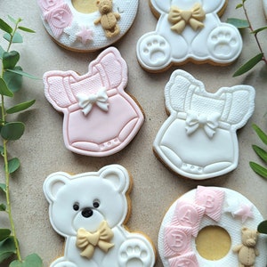 May include: Six decorated sugar cookies for a baby shower. Two cookies are shaped like teddy bears with bows, two are shaped like baby onesies, and two are shaped like a donut with the word "BABY" on them.