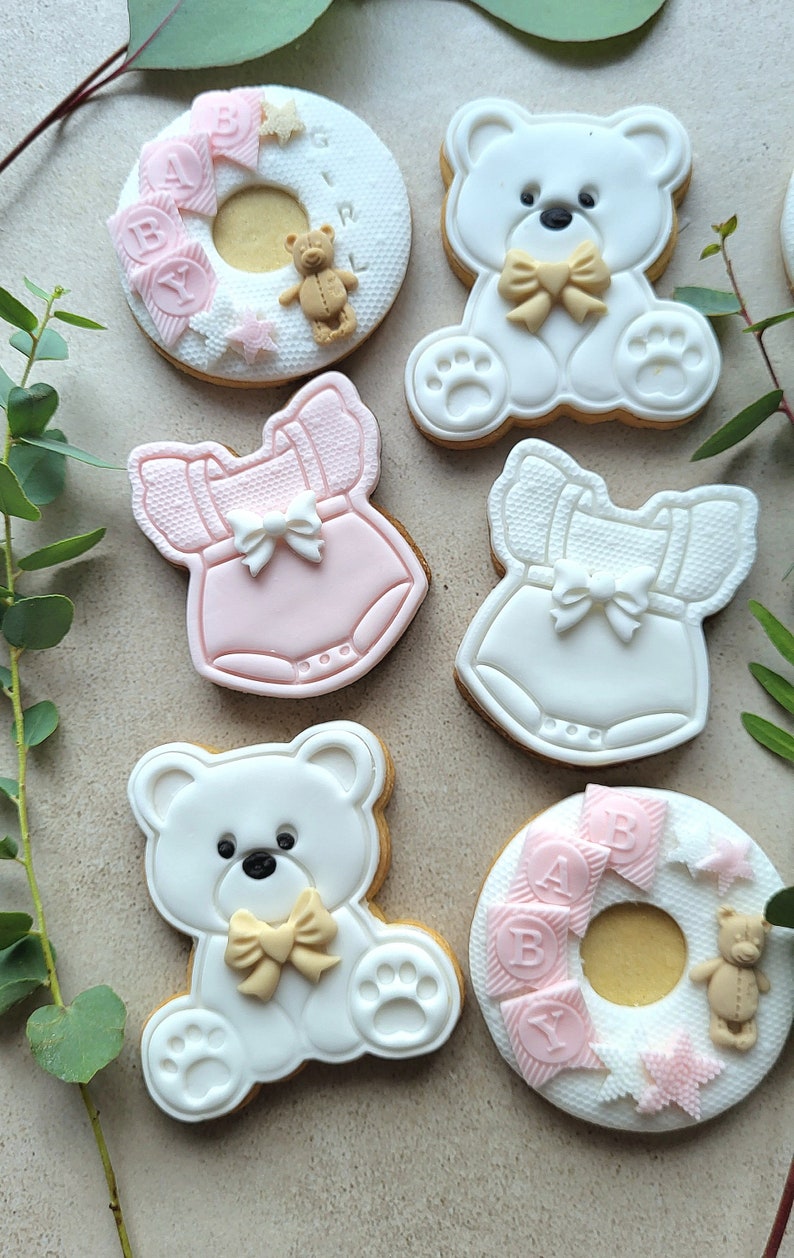 May include: Six decorated sugar cookies for a baby shower. Two cookies are shaped like teddy bears with bows, two are shaped like baby onesies, and two are shaped like a donut with the word "BABY" on them.