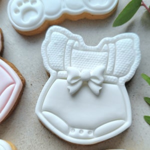 May include: A set of white sugar cookies decorated with fondant. One cookie is shaped like a baby onesie with a white bow, another is shaped like a teddy bear with a yellow bow, and another is shaped like a block with the letter "B" on it.