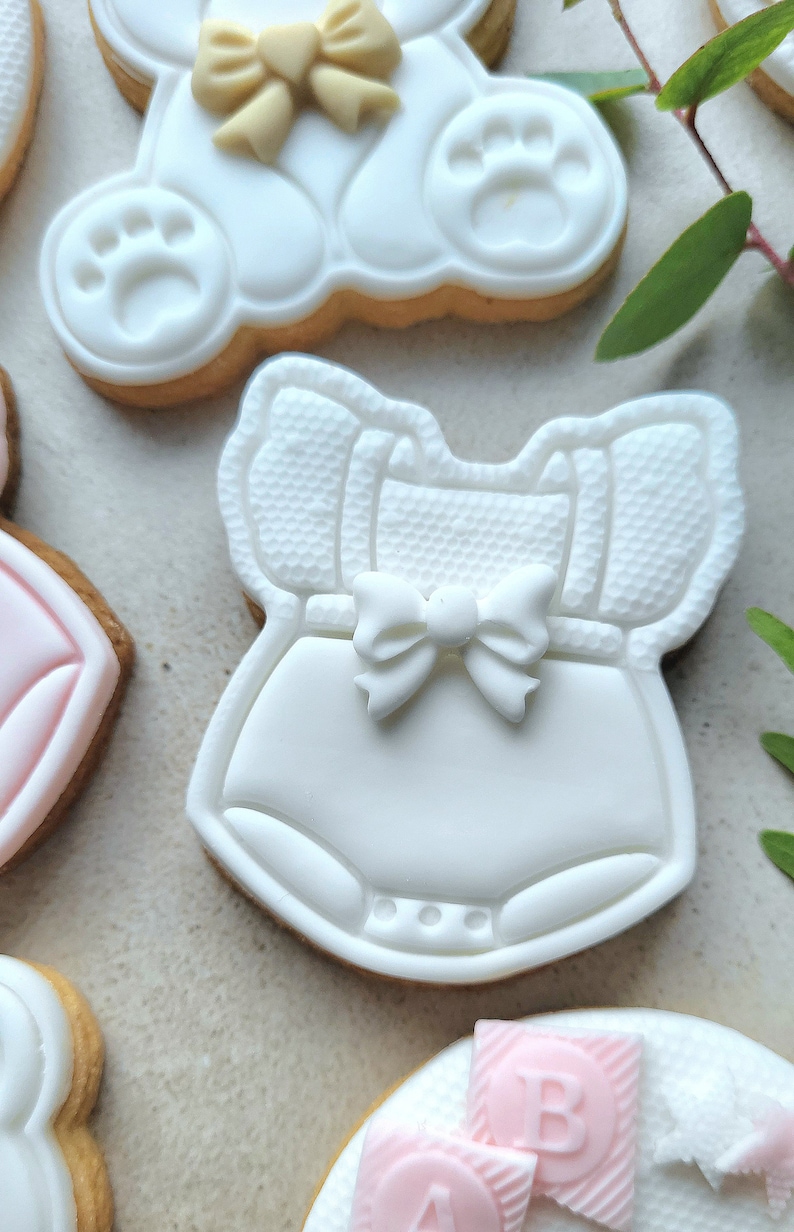 May include: A set of white sugar cookies decorated with fondant. One cookie is shaped like a baby onesie with a white bow, another is shaped like a teddy bear with a yellow bow, and another is shaped like a block with the letter "B" on it.