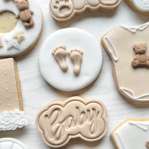 May include: A set of nine decorated sugar cookies with a baby theme. The cookies feature a teddy bear, a baby onesie, a baby carriage, footprints, and the word "Baby".