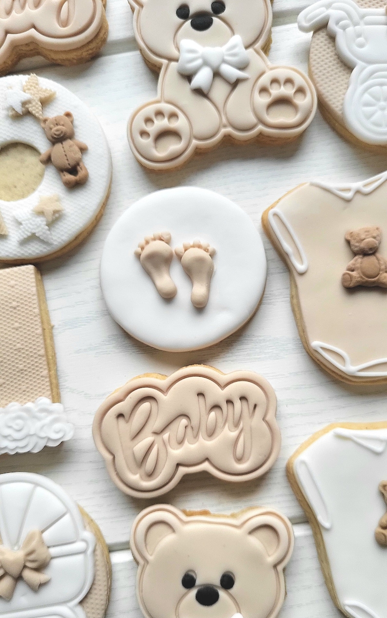 May include: A set of nine decorated sugar cookies with a baby theme. The cookies feature a teddy bear, a baby onesie, a baby carriage, footprints, and the word "Baby".