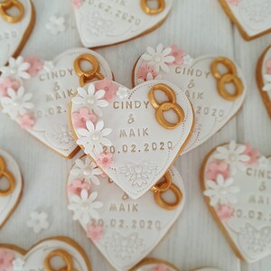 May include: Heart-shaped cookies decorated with white icing, pink flowers, and gold rings. The cookies have the text "Cindy & Maik" and the date "20.02.2020" written on them.