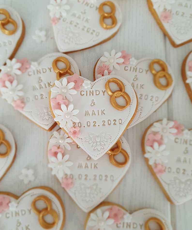 May include: Heart-shaped cookies decorated with white icing, pink flowers, and gold rings. The cookies have the text "Cindy & Maik" and the date "20.02.2020" written on them.