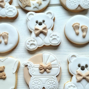 May include: A set of nine decorated sugar cookies featuring a baby theme. The cookies are decorated with white icing and brown accents. Some cookies feature teddy bears, a baby carriage, and footprints.