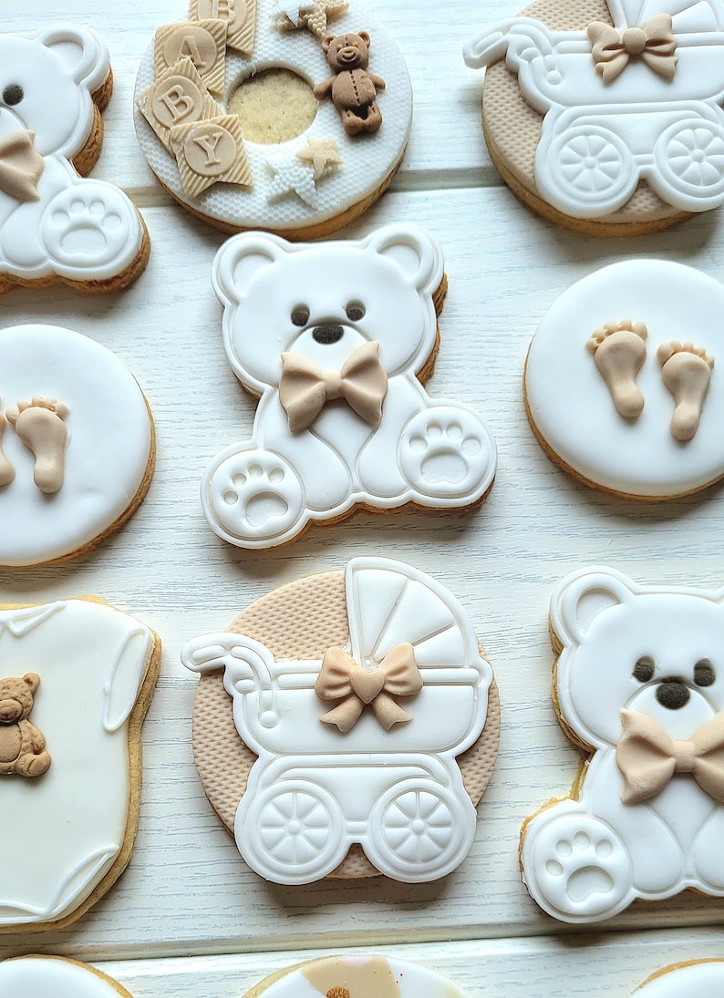 May include: A set of nine decorated sugar cookies featuring a baby theme. The cookies are decorated with white icing and brown accents. Some cookies feature teddy bears, a baby carriage, and footprints.