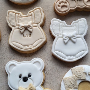 May include: Assortment of six decorated sugar cookies. The cookies are decorated with white and brown icing. The designs include a teddy bear, a baby onesie with the name "LIU" on it, and a ring of baby blocks that spell "BABY".