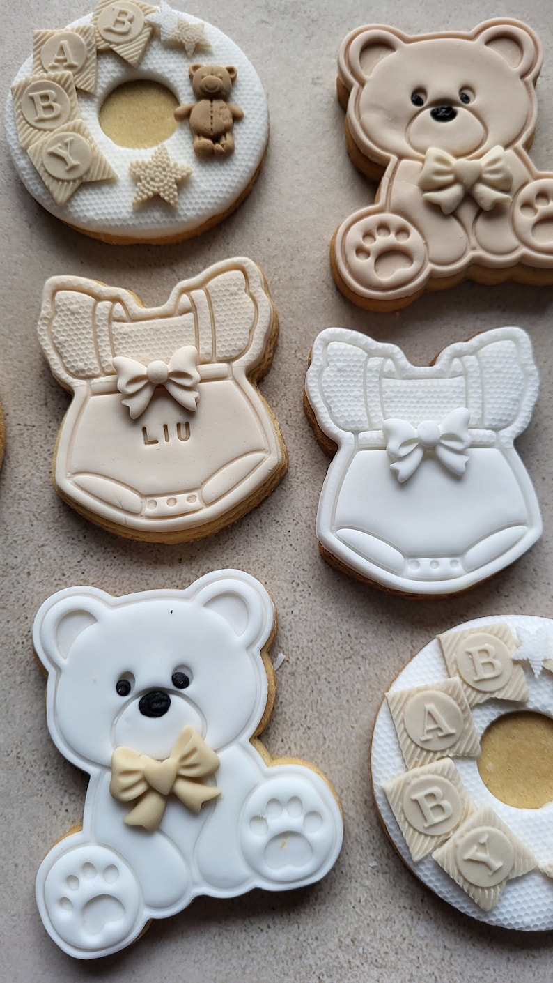 May include: Assortment of six decorated sugar cookies. The cookies are decorated with white and brown icing. The designs include a teddy bear, a baby onesie with the name "LIU" on it, and a ring of baby blocks that spell "BABY".