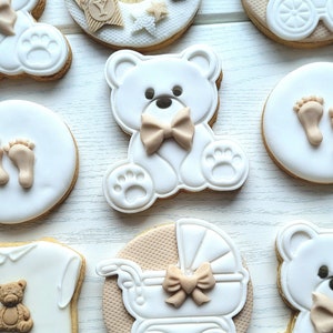 May include: A set of nine decorated sugar cookies featuring a baby theme. The cookies are decorated with white icing and brown accents. The designs include a teddy bear, a baby carriage, a baby footprint, and a baby onesie.