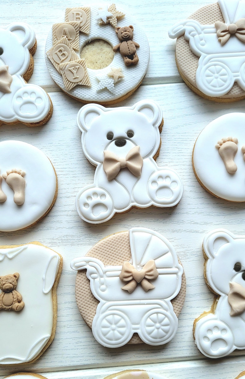 May include: A set of nine decorated sugar cookies featuring a baby theme. The cookies are decorated with white icing and brown accents. The designs include a teddy bear, a baby carriage, a baby footprint, and a baby onesie.