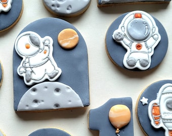 May include: Assortment of space-themed cookies with gray, white, and orange icing. Designs include astronauts, planets, moons, and the text "Happy Space." One cookie is shaped like the number one with a balloon and the name "Liam."