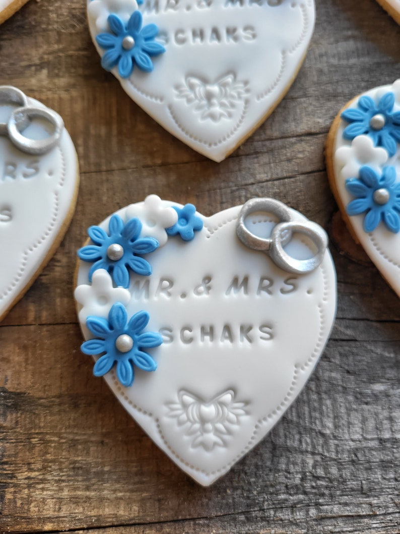 May include: White heart-shaped cookies decorated with blue flowers, silver rings, and the text "MR. & MRS. SCHAKS".