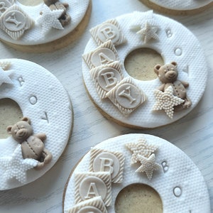 May include: Four decorated sugar cookies with white icing and brown teddy bears. The cookies are shaped like a ring and have the words "Baby Boy" written on them. The cookies are decorated with brown blocks that spell out "Baby" and white stars.