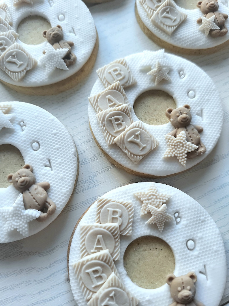 May include: Four decorated sugar cookies with white icing and brown teddy bears. The cookies are shaped like a ring and have the words "Baby Boy" written on them. The cookies are decorated with brown blocks that spell out "Baby" and white stars.