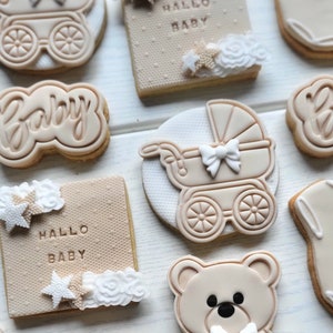 May include: A set of nine decorated sugar cookies with a baby theme. The cookies feature a teddy bear, a baby carriage, a star, and the words "Hallo Baby" and "Baby".