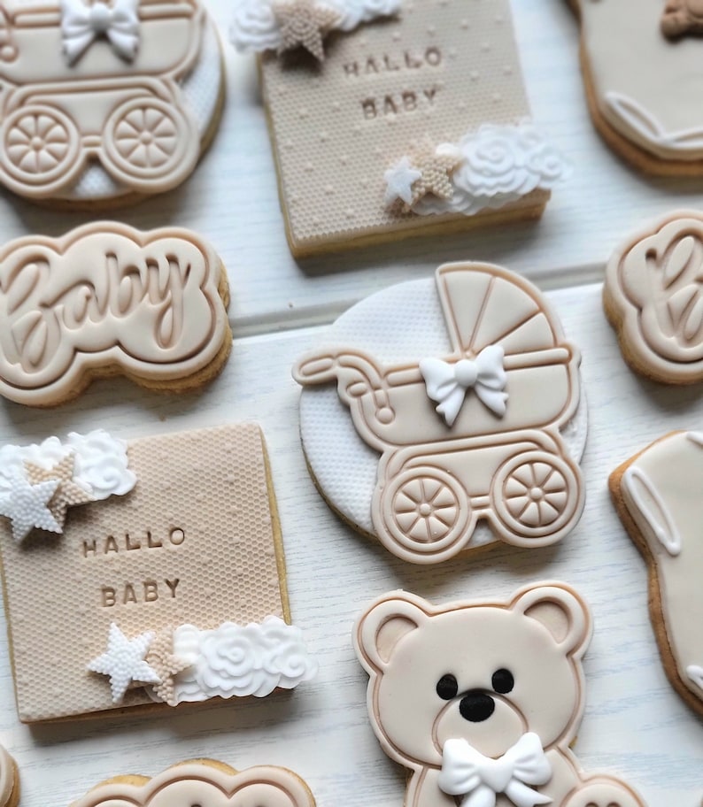 May include: A set of nine decorated sugar cookies with a baby theme. The cookies feature a teddy bear, a baby carriage, a star, and the words "Hallo Baby" and "Baby".
