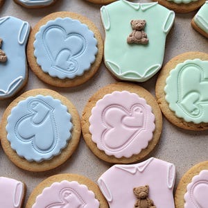 May include: An assortment of baby-themed cookies. Some are shaped like onesies with a small teddy bear, others are round with heart designs. Colors include blue, pink, and green, with a light brown cookie base.