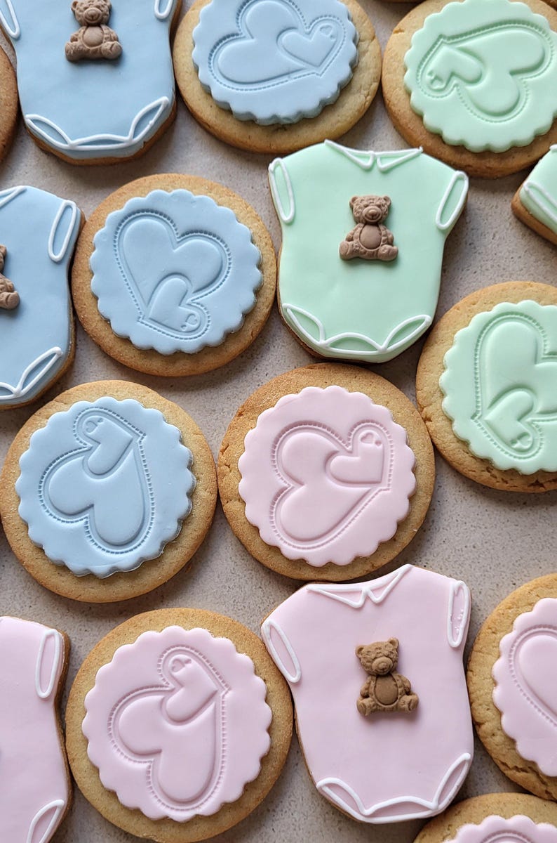 May include: An assortment of baby-themed cookies. Some are shaped like onesies with a small teddy bear, others are round with heart designs. Colors include blue, pink, and green, with a light brown cookie base.