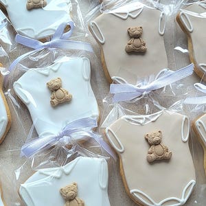 May include: Cookies shaped like baby onesies, decorated with a teddy bear and tied with a light blue ribbon. Some cookies are white, others are tan, all are individually wrapped in clear cellophane.