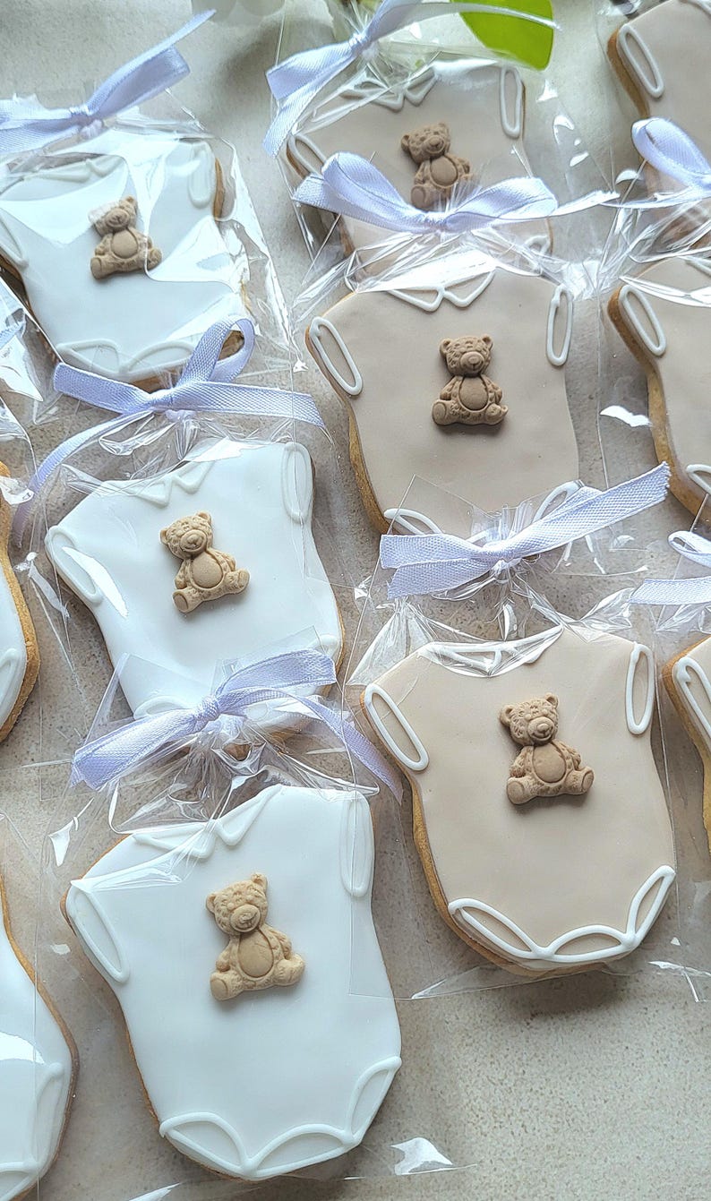 May include: Cookies shaped like baby onesies, decorated with a teddy bear and tied with a light blue ribbon. Some cookies are white, others are tan, all are individually wrapped in clear cellophane.