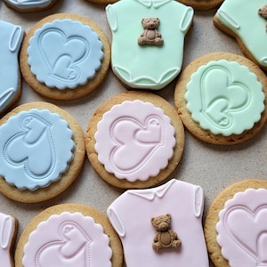May include: Assortment of baby-themed cookies. Some cookies are round with heart designs in blue, pink, and green. Others are shaped like baby onesies, also in pastel colors, and topped with small teddy bear decorations. The cookies are on a light-colored surface.
