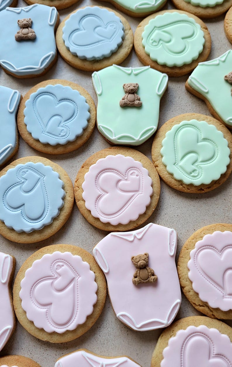 May include: Assortment of baby-themed cookies. Some cookies are round with heart designs in blue, pink, and green. Others are shaped like baby onesies, also in pastel colors, and topped with small teddy bear decorations. The cookies are on a light-colored surface.
