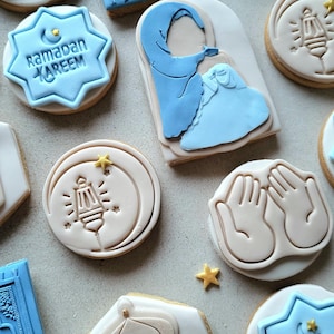Peut inclure: Assortiment de biscuits sur le thème du Ramadan avec glaçage bleu et beige. Les motifs comprennent une mosquée, un tapis de prière, des mains en prière et les mots "Ramadan Kareem" dans une forme d'étoile. Des étoiles dorées sont également présentes.