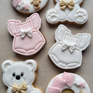 May include: Six decorated sugar cookies, two are shaped like a teddy bear with a bow, two are shaped like a baby onesie, and two are shaped like a ring with the word "BABY" on it.
