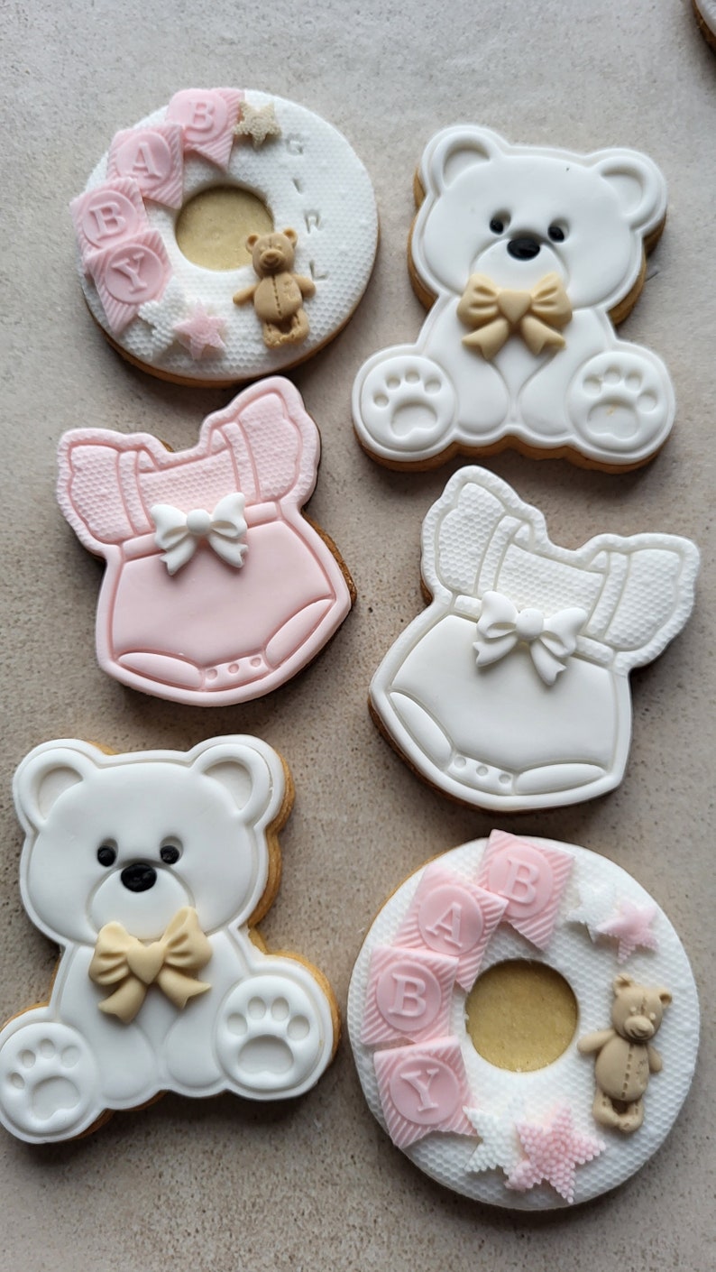 May include: Six decorated sugar cookies, two are shaped like a teddy bear with a bow, two are shaped like a baby onesie, and two are shaped like a ring with the word "BABY" on it.