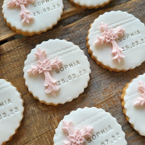 May include: Round cookies with white icing and pink cross decorations. Each cookie has the name "SOPHIA" and a date. The cookies are arranged on a wooden surface, with a scalloped edge.