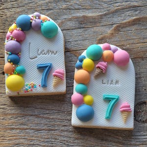 Könnte beinhalten: Zwei dekorierte Zuckerkekse mit einem Bogen aus bunten Luftballons, Eiswaffeln und dem Namen Liam und der Zahl 7 auf jedem Keks.