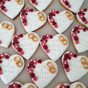 May include: A collection of heart-shaped cookies decorated with white icing, gold rings, and red flowers. The cookies are decorated with the text "EVIN & NECDET 01.02.20".