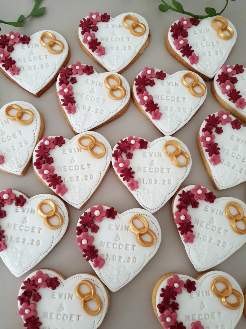 May include: A collection of heart-shaped cookies decorated with white icing, gold rings, and red flowers. The cookies are decorated with the text "EVIN & NECDET 01.02.20".