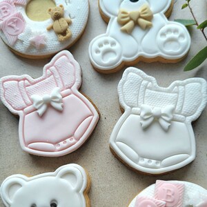 May include: Assortment of decorated sugar cookies for a baby shower. The cookies feature a pink and white baby onesie, a white teddy bear, and a ring-shaped cookie with the word "Baby" in pink letters.