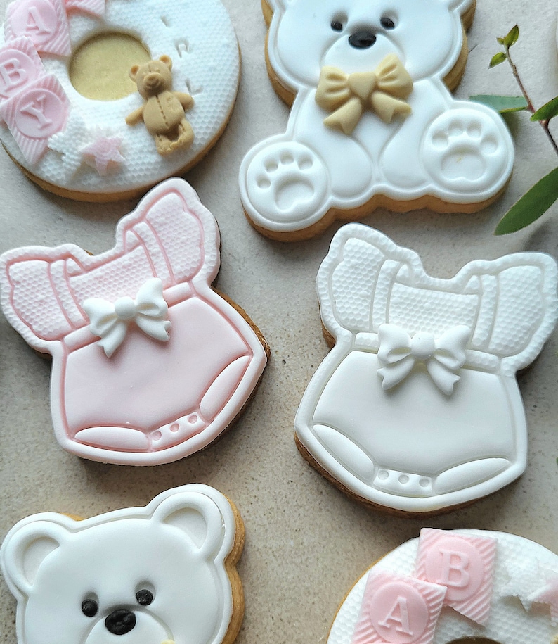May include: Assortment of decorated sugar cookies for a baby shower. The cookies feature a pink and white baby onesie, a white teddy bear, and a ring-shaped cookie with the word "Baby" in pink letters.