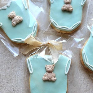 May include: Baby shower cookies shaped like onesies, iced in light blue with white trim. Each cookie has a small teddy bear decoration. Cookies are individually wrapped and tied with a gold ribbon.