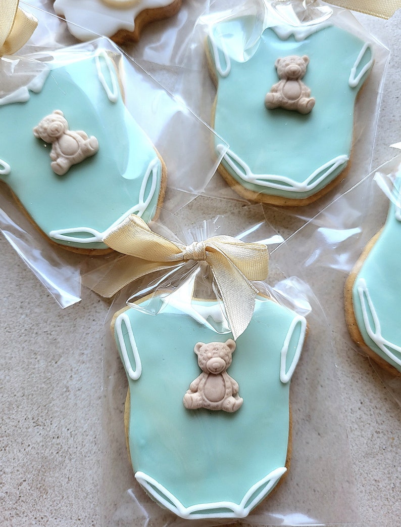 May include: Baby shower cookies shaped like onesies, iced in light blue with white trim. Each cookie has a small teddy bear decoration. Cookies are individually wrapped and tied with a gold ribbon.