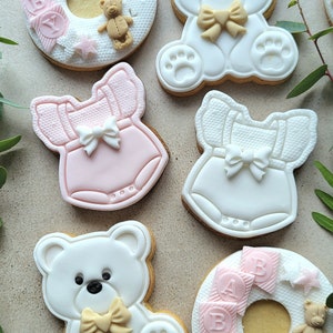May include: Six decorated sugar cookies, including two teddy bear cookies, two cookies shaped like baby onesies, and two ring-shaped cookies with the words "BABY" and "GIRL" in pink letters.