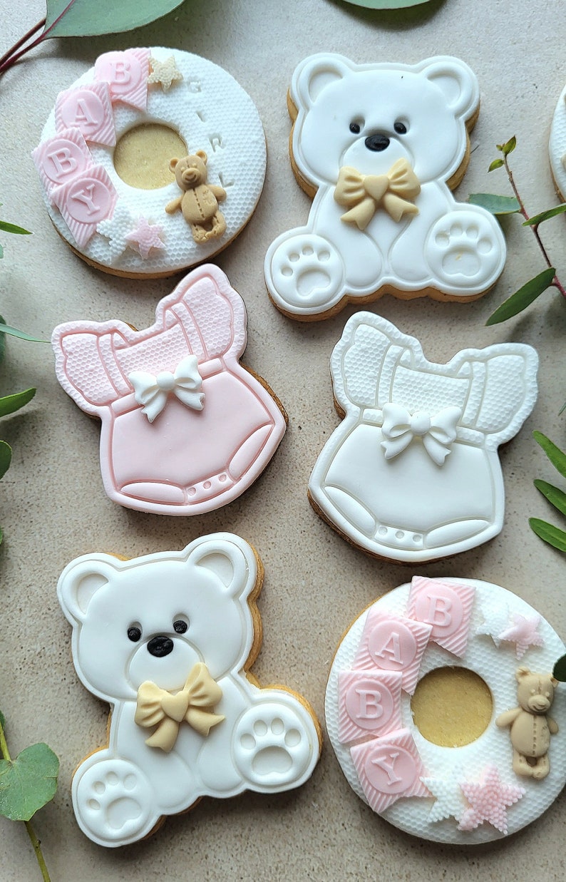 May include: Six decorated sugar cookies, including two teddy bear cookies, two cookies shaped like baby onesies, and two ring-shaped cookies with the words "BABY" and "GIRL" in pink letters.