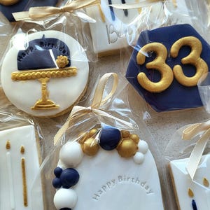 May include: A collection of individually wrapped sugar biscuits featuring navy blue and white icing with gold accents. Designs include the number 33, birthday candles, a cake on a stand, and an arch with balloons and the text Happy Birthday Benjamin.